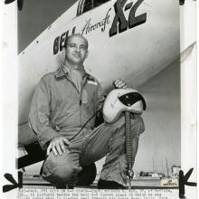 A man kneels in a flight suit in front of the nose of an aircraft which has Bell Aircraft X-2 written on the side. He balances a helmet on his knee.