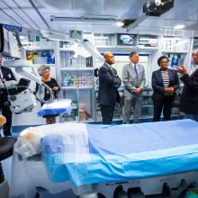 A group of people stand in a semi circle talking inside an operating room.