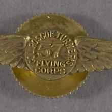 A gold pin in the shape of wings with the number 57 at the center. Around the 57 it reads Col. Roscoe Turner's Flying Corps.