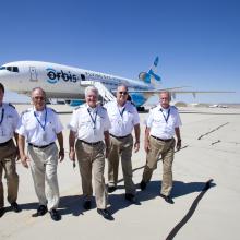 Five pilots walking toward the camera shoulder to shoulder. The Orbis Flying Eye Hospital can be see in the background.