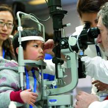 A small child looks into optometry equipment while a doctor takes measurements.