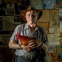 A man faces the camera holding a chicken. Behind him are various maps and newspapers hanging on the wall.