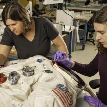 Two women next to a spacesuit working to conserve it with tools.