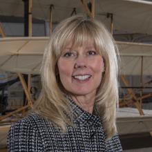 A blonde woman smiles at the camera with a plane behind her. 