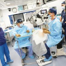 Four medical professionals stand around an operating table. One of them sits next to the table looking into a machine that is focuses on the patient.