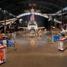 At the center of the photograph is the nose of the Space Shuttle, with the Space Shuttle expanding behind it. Hanging overhead is a satellite, a spacesuit, and a missile. Surrounding on the ground are cases with space stuff. 