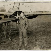 A man in a flying suit stands in front of an aircraft in a field. 