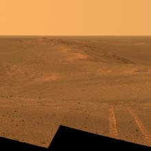 An artist's rendering of a dusty red landscape with hills or dunes. A set of two tire marks appears at the lower right side of the screen. 