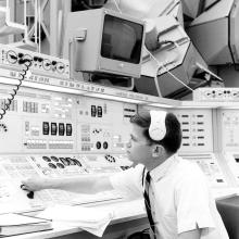 A man with a headset sits at a set of controls and computers. He appears to be flipping the position of a knob.