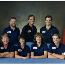 Astronaut group portrait wearing navy polo shirts with NASA logo on right breast--three men standing behind two men with two women seated between them.  American flag on pole to the left, NASA flag on pole to the right. On the table: replica of gold astronaut pin on left, model of the space shuttle on the right.