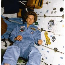 Woman in a light blue NASA flight suit floats weightlessly in front of the white lockered walls of the Space Shuttle.