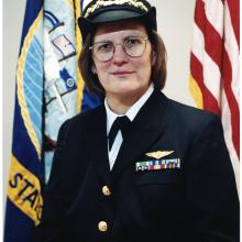 Portrait of a woman wearing glasses in a naval uniform with yellow stripes at the ends of the sleeves.  Her cap has a white top and black brim with a Navy insignia.  Behind her are the Navy flag and US flag.