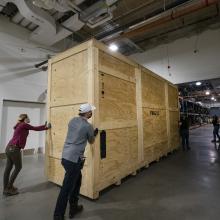 Staff members push a large wooden crate through a large hangar setting.