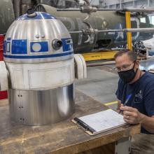 Man sits at a table writing on a pad of paper next to the top half of a model of R2-D2, a silver and blue droid.