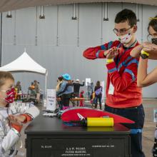 Two young adults wearing costumes demonstrate the wing of an aircraft with their hands. A young child in front of them mimics their hand motions.