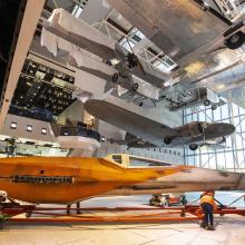 A group of workers in construction vests and hats move the fuselage of the X-wng, a pointed orange and white spacecraft, through a nearly-empty gallery with aircraft hanging above.