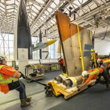 A group of people in orange construction vest and hat push the wing of an X-wing down the hall of a museum with rockets in the background.