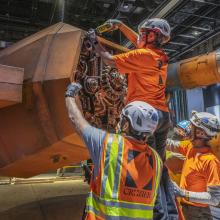 Four workers in construction vests and helmets assemble the engines of a Star War X-wing, an orange flying vehicle.