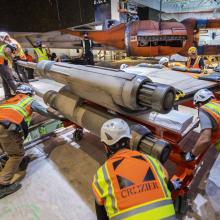 A group of workers in orange construction vests and helmets move part of the X-wing in the construction area at the Museum in DC.