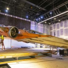 A cream an orange full-size X-wing hanging a few feet over the ground as it is raised.