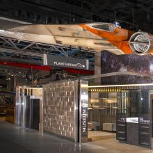 X-wing, a weathered cream and orange space vehicle hangs over an unstocked store and ticket area.