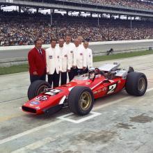 One man in a red jacket and six med in white jackets standing behind a red race car.