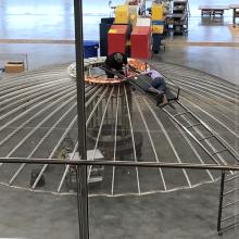 Two individuals work on the top of an umbrella shaped satellite in a restoration hangar.