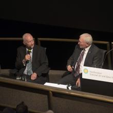 Two men sitting in chair with business suits on and microphones in their hands.