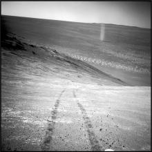 Two wheel tracks on the surface of Mars are in the foreground. In the background is a structure forming that looks similar to a tornado.