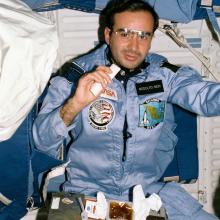 An astronaut in a blue suit floats inside a spacecraft. His food tray is on his lap, a tortilla in his hand. 