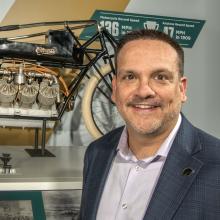 A man in a blazer smiles in front of the camera in front of a Curtiss bicycle. 