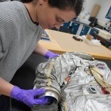 A woman wearing purple gloves holds a silver spacesuit. 