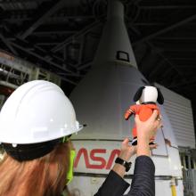 A woman wearing a hard-hat, holds a stuffed animal Snoopy facing the top of the SLS rocket.