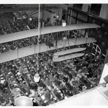 A photo taken from a balcony looking down at a crowd of seated people. The Wright Flyer hangs overs head, with the Spirit of St. Louis hung across from it.