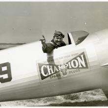 Side view of Roscoe Turner seated in the cockpit of his LTR-14 "Meteor" airplane on the ground. Turner points his right hand and winks.