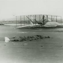 A black and white photo of a biplane on a sandy beach with a man standing the the far left. The front elevator is almost perpendicular to the plane as it is damaged.