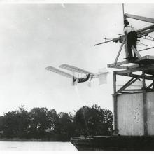 One-half left rear view of a Langley Aerodrome No. 5 in flight over the Potomac River just after launch. A man watches from a houseboat at top right.