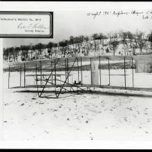 The Wright Flyer in the foreground of a snowy field, seen from the front. There is an exhibit number stamped at the top left.