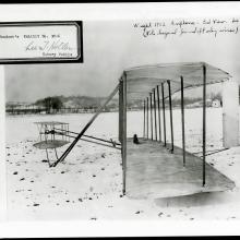 The Wright Flyer as seen from the side in a snowy field. There is an exhibit number stamped at the top left.