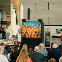 Astronauts are shown on a small tv screen while a small ground looks towards it.