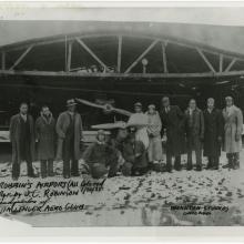 A group of eleven people stand in a line next to each other in front of an aircraft hangar. An airplane can be seen directly behind the group.