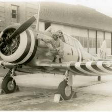 Sepia toned image of Jesse Leroy Brown squatting on the right side wing of an aircraft as he poses for a picture.