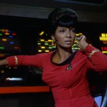 A Black woman in a red starfleet uniform looks up from a dashboard. 