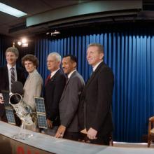 A group of 5 people stand shoulder to shoulder in front of a model of a telescope behind a table. On the other side of the table are members of the press.