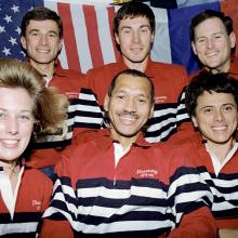 Six people pose in two rows of three aboard a space shuttle. The woman in the left demonstrates the weightlessness of space with her hair floating freely.