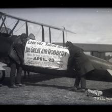 Men affix banner to side of biplane fuselage which reads; Look Out Staten Island / The Aerial Mail Bandits in / The Great Air Robbery / Will Bomb You From The Sky / April 23. If You Hear A Curtis [sic] Aeroplane / Don't Look Up.