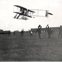 A black and white image of a biplane flying low to the ground. People on the ground wave their caps. 