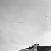 Distant right rear view of a Pitcairn PCA-2 Autogiro towing an aerial banner reading, "Adler Shoes for Men - sized 4 to 14 & 24."Due to the direction of flight, the banner reads in reverse.