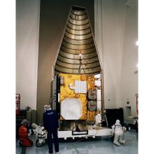 A large golden colored cube inside a metal conical encasing is surrounded by various technicians performing different activities on the structure.