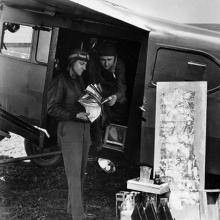 Two men stand in the door of a plane looking at photography equipment. 